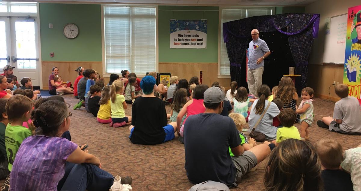 Picture of large meeting room with families seated on the floor and a standing performer.